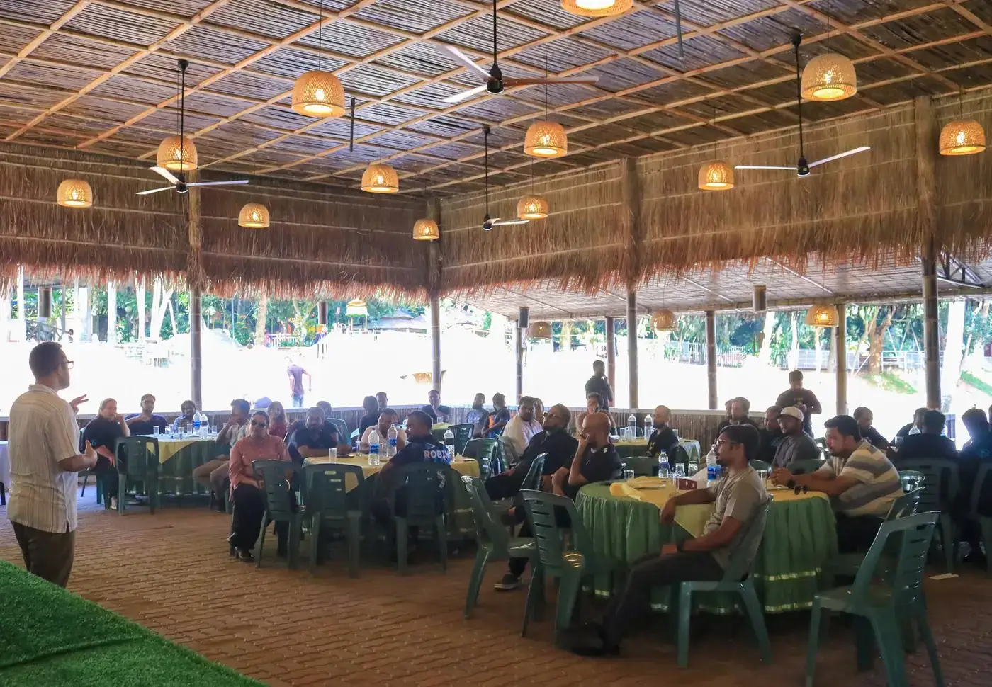 A facilitator speaking to participants seated at round tables in an open-air thatched hall